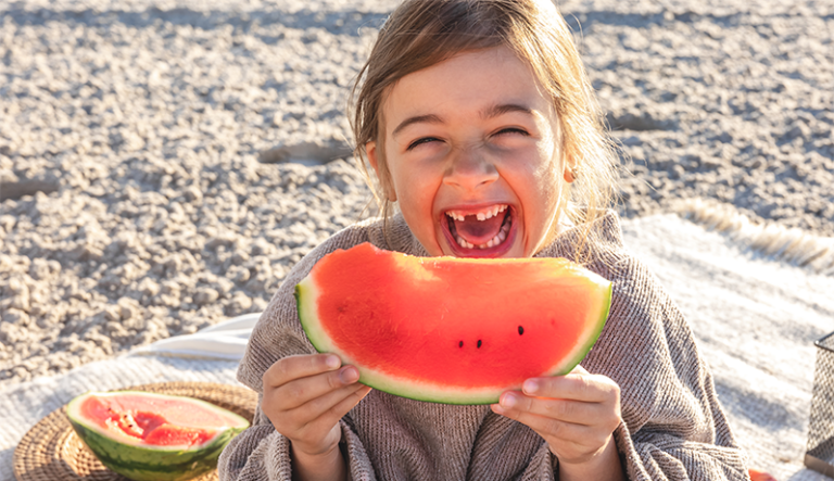 Obst_psychische-Gesundheit.png Ein lachendes Kind hält ein großes Stück Wassermelone in der Hand, umgeben von einer sommerlichen Picknick-Atmosphäre.