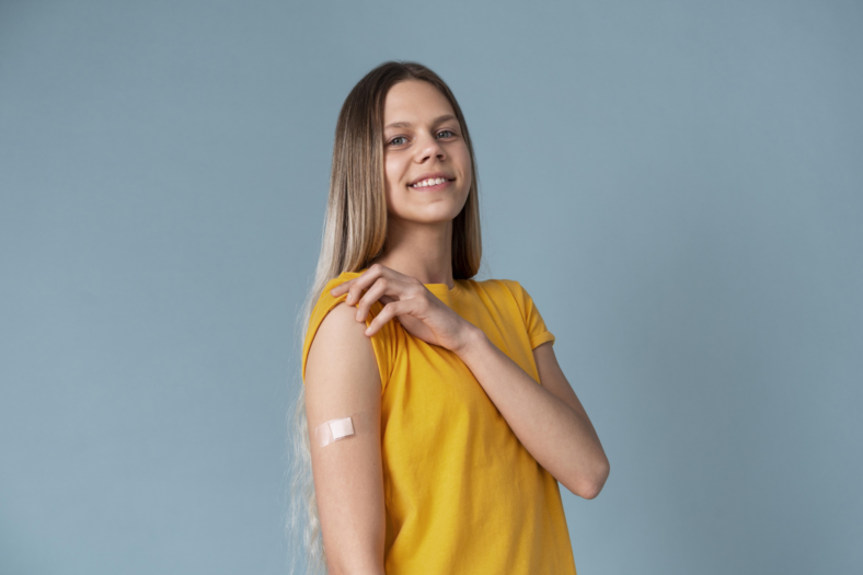 HPV_Impfung.png Eine lächelnde junge Frau mit langen Haaren trägt ein gelbes T-Shirt und zeigt einen Verband am Oberarm.